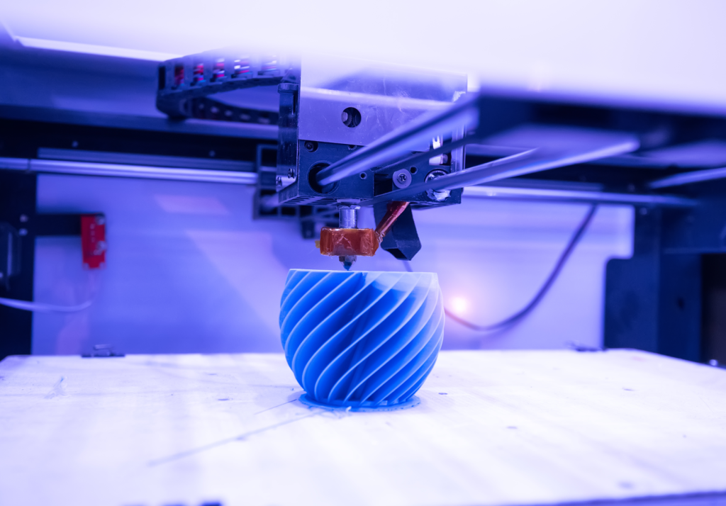 Detal w kolorze niebieskim o spiralnym kształcei powierzchni bocznej, w trakcie procesu drukowania 3D, znajdujący się na stole roboczym drukarki, pod głowicą drukującą