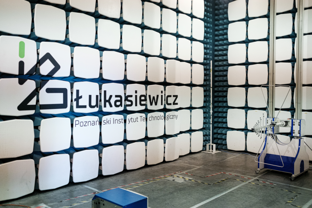 Komora bezechowa z białymi panelami akustycznymi na ścianie, z logo i napisem 'Łukasiewicz Poznański Instytut Technologiczny', wyposażona w sprzęt pomiarowy