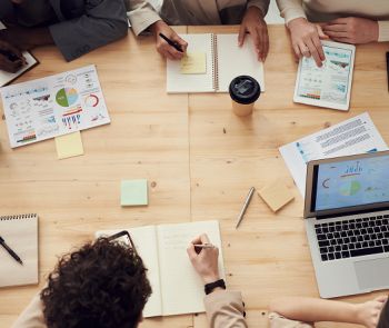 Blat stołu z rozłożonymi na nim notatnikami, komputerami i przyborami do pisania. Wokół stołu siedzą pracownicy.