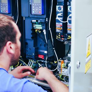 osoba sprawdzająca układ elektryczny pojazdu szynowego, w tle rozdzielnia, kable i bezpieczniki